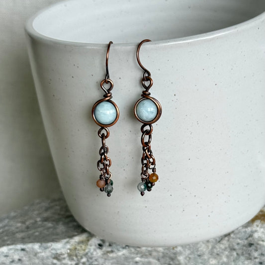 Pair of copper earrings with blue gemstones and copper accents hanging over a white ceramic bowl.
