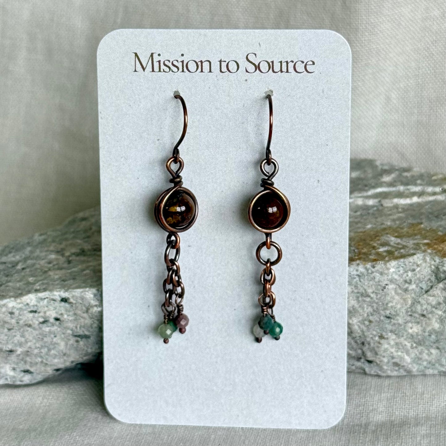 Pair of copper earrings on a 'Mission to Source' card with rocks in the background