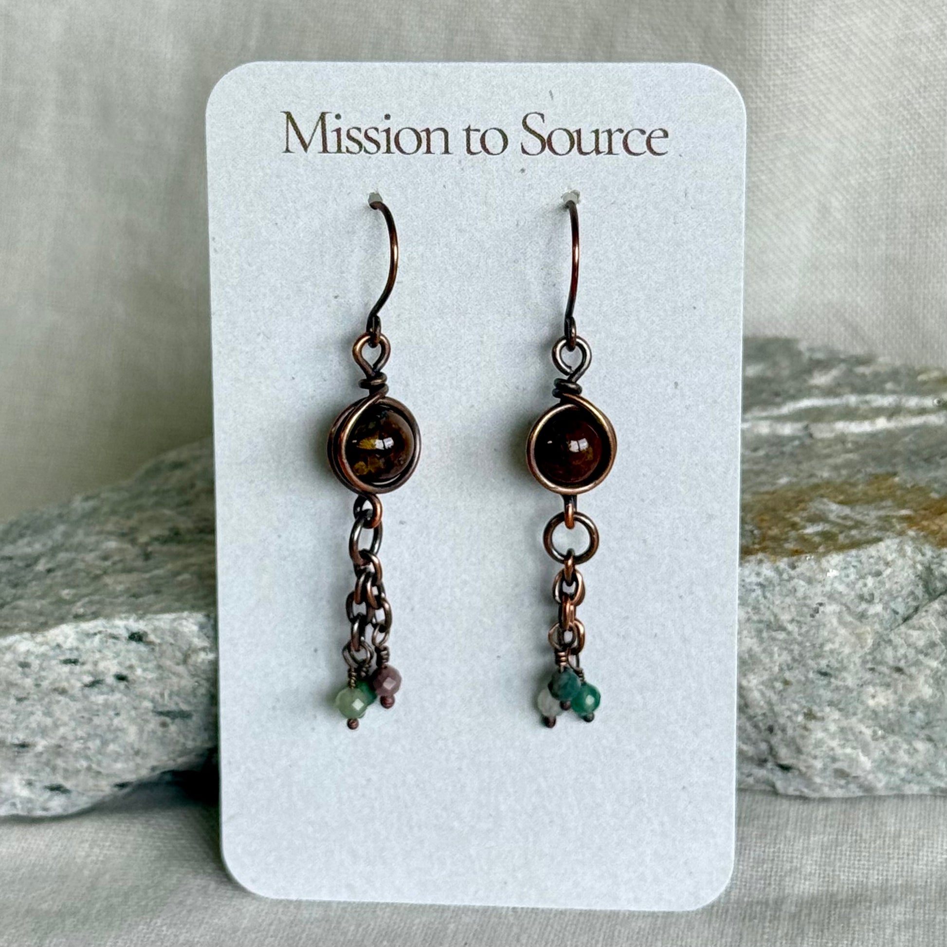 Pair of copper earrings on a 'Mission to Source' card with rocks in the background