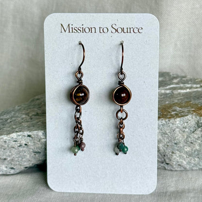 Pair of copper earrings on a 'Mission to Source' card with rocks in the background