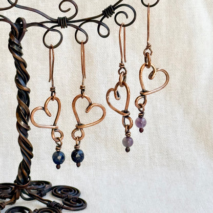 Heart-shaped copper earrings with beads on a decorative stand against a beige background