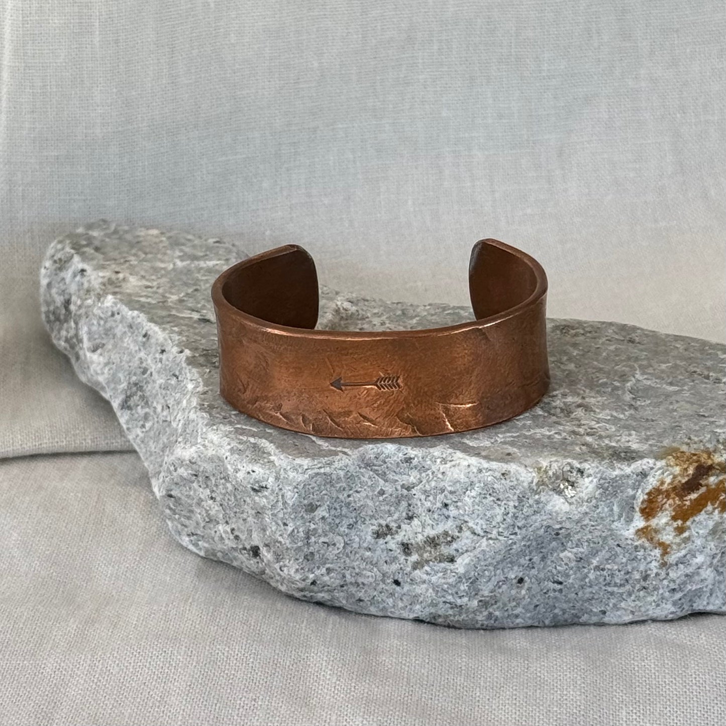 Copper cuff with an embossed  arrow design on a stone surface with linen background