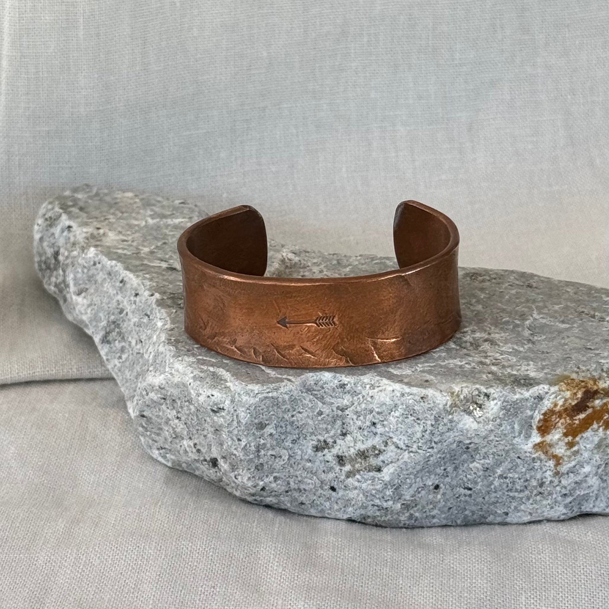 Copper cuff with an embossed  arrow design on a stone surface with linen background