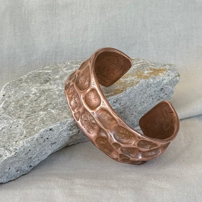 Copper cuff with hammered, textured design leaning on a stone surface with linen background