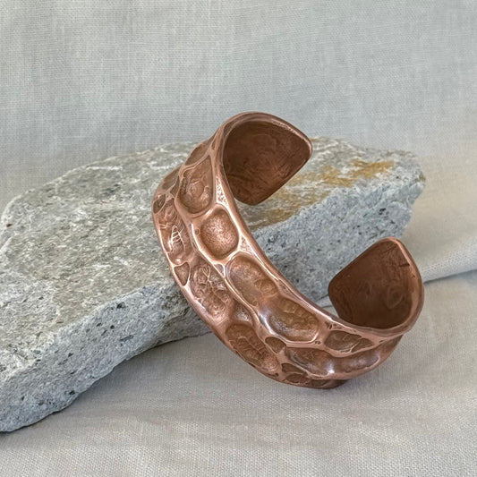 Copper cuff with hammered, textured design leaning on a stone surface with linen background
