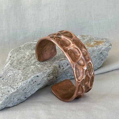 Copper cuff with hammered, textured design on a stone surface with linen background