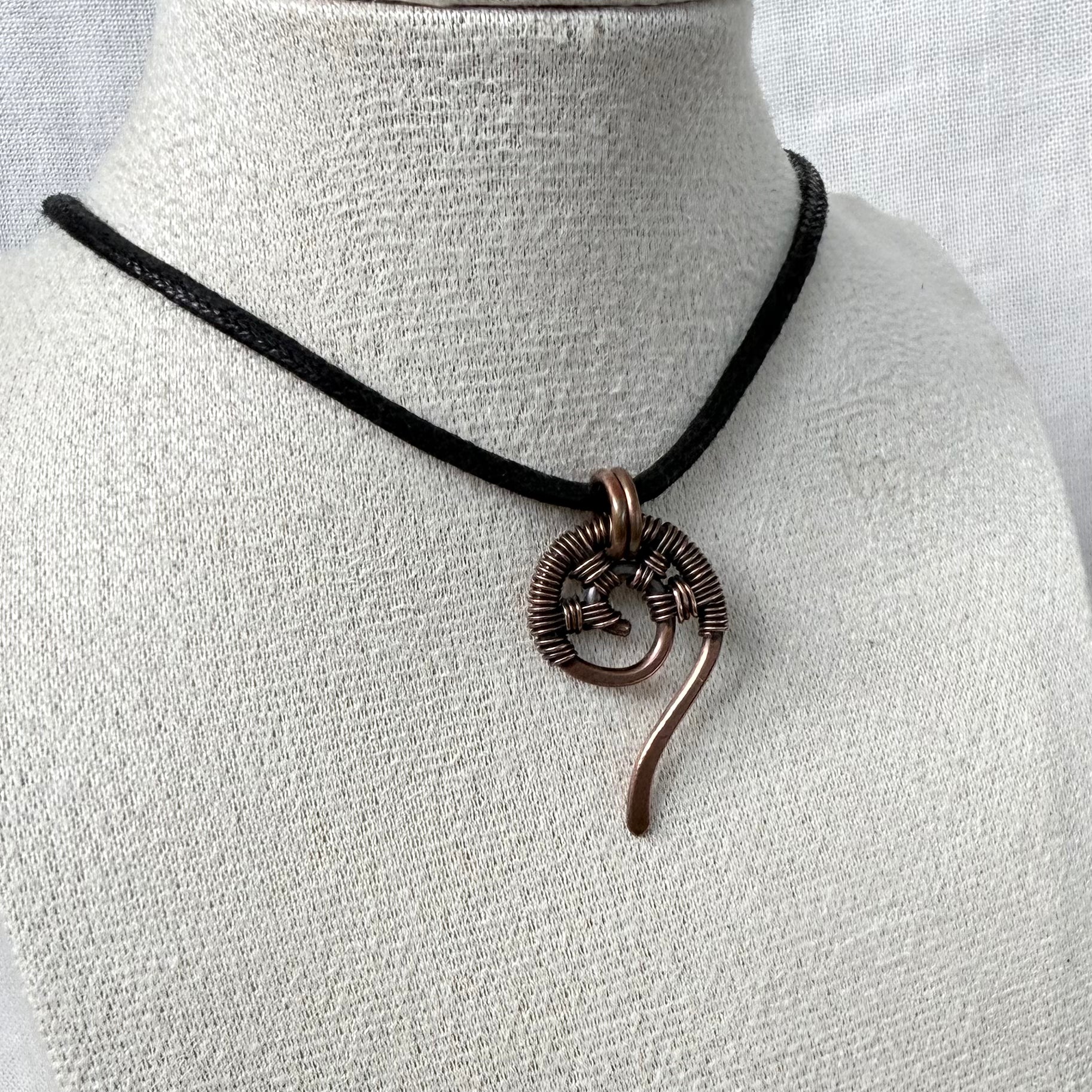 A handmade necklace featuring a textured copper spiral pendant on a black cord, displayed on a textured white fabric mannequin bust.