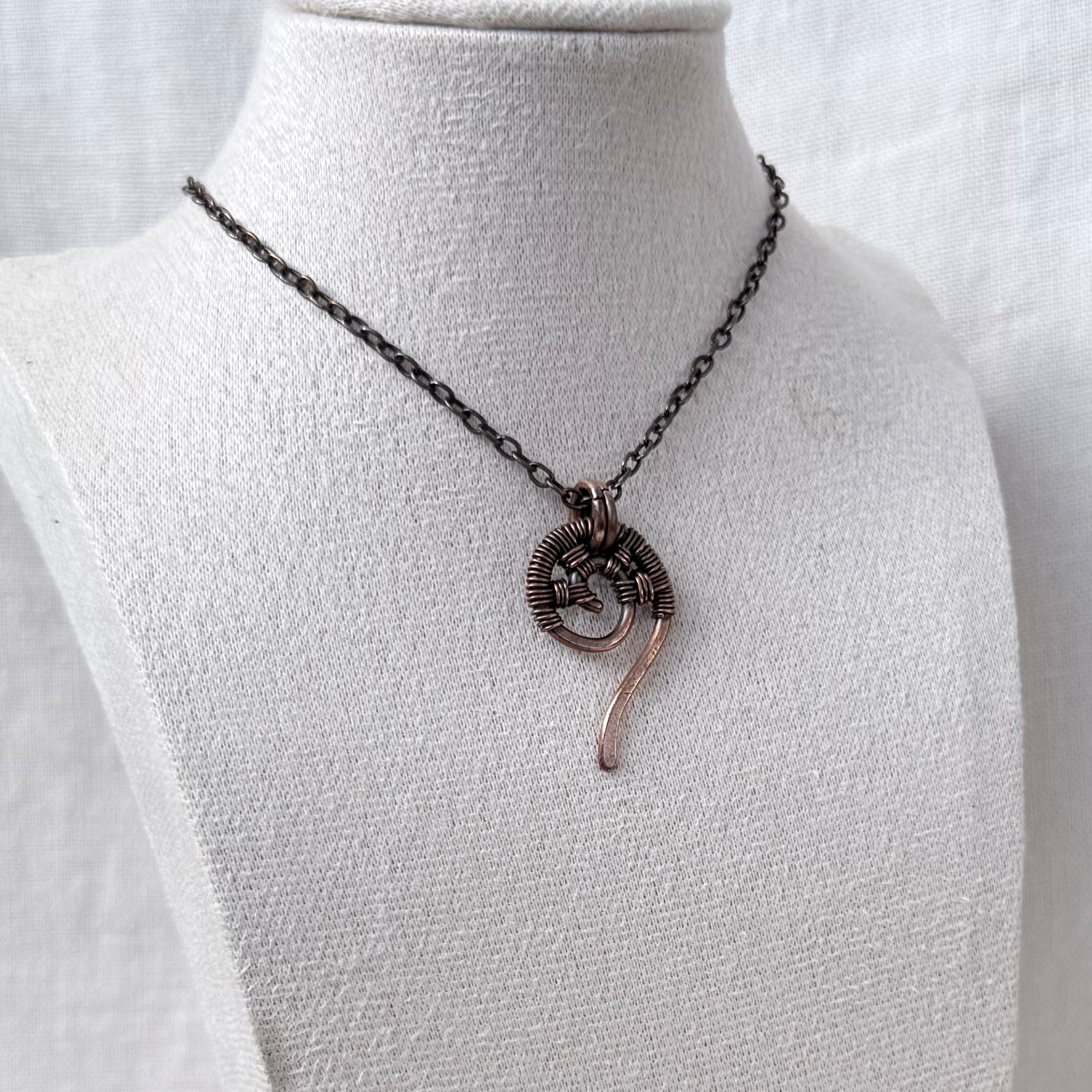A handmade copper wire-wrapped necklace with a spiral pendant and a dangling curved accent, displayed on a white fabric jewelry bust.