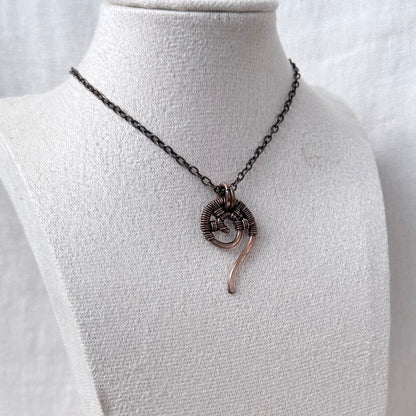 A handmade copper wire-wrapped necklace with a spiral pendant and a dangling curved accent, displayed on a white fabric jewelry bust.