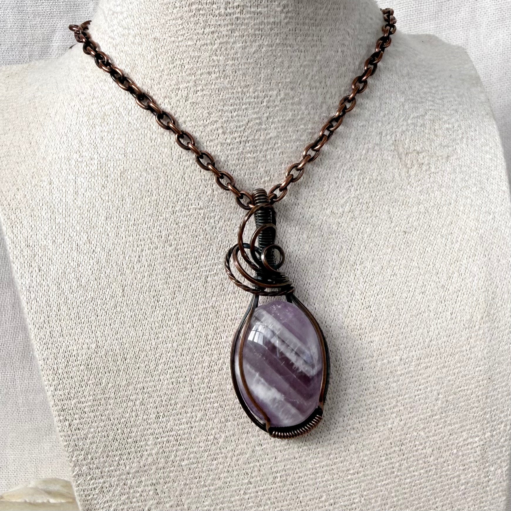 A teardrop-shaped purple and white banded Amethyst stone pendant wrapped in dark copper wire, hanging from a dark copper chain on a textured off-white fabric display bust.