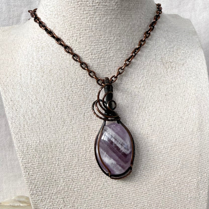 A teardrop-shaped purple and white banded Amethyst stone pendant wrapped in dark copper wire, hanging from a dark copper chain on a textured off-white fabric display bust.