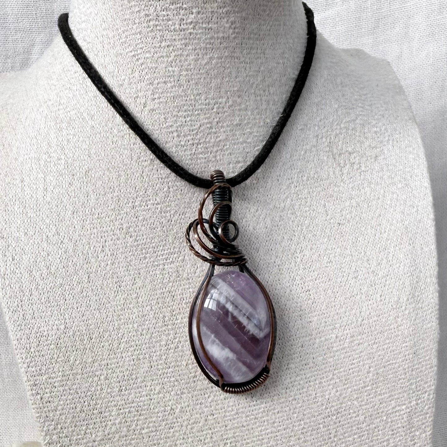 A teardrop-shaped purple Amethyst pendant wrapped in dark copper wire, hanging from a black cord necklace on a textured white mannequin bust.