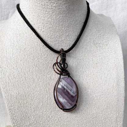 A teardrop-shaped purple Amethyst pendant wrapped in dark copper wire, hanging from a black cord necklace on a textured white mannequin bust.