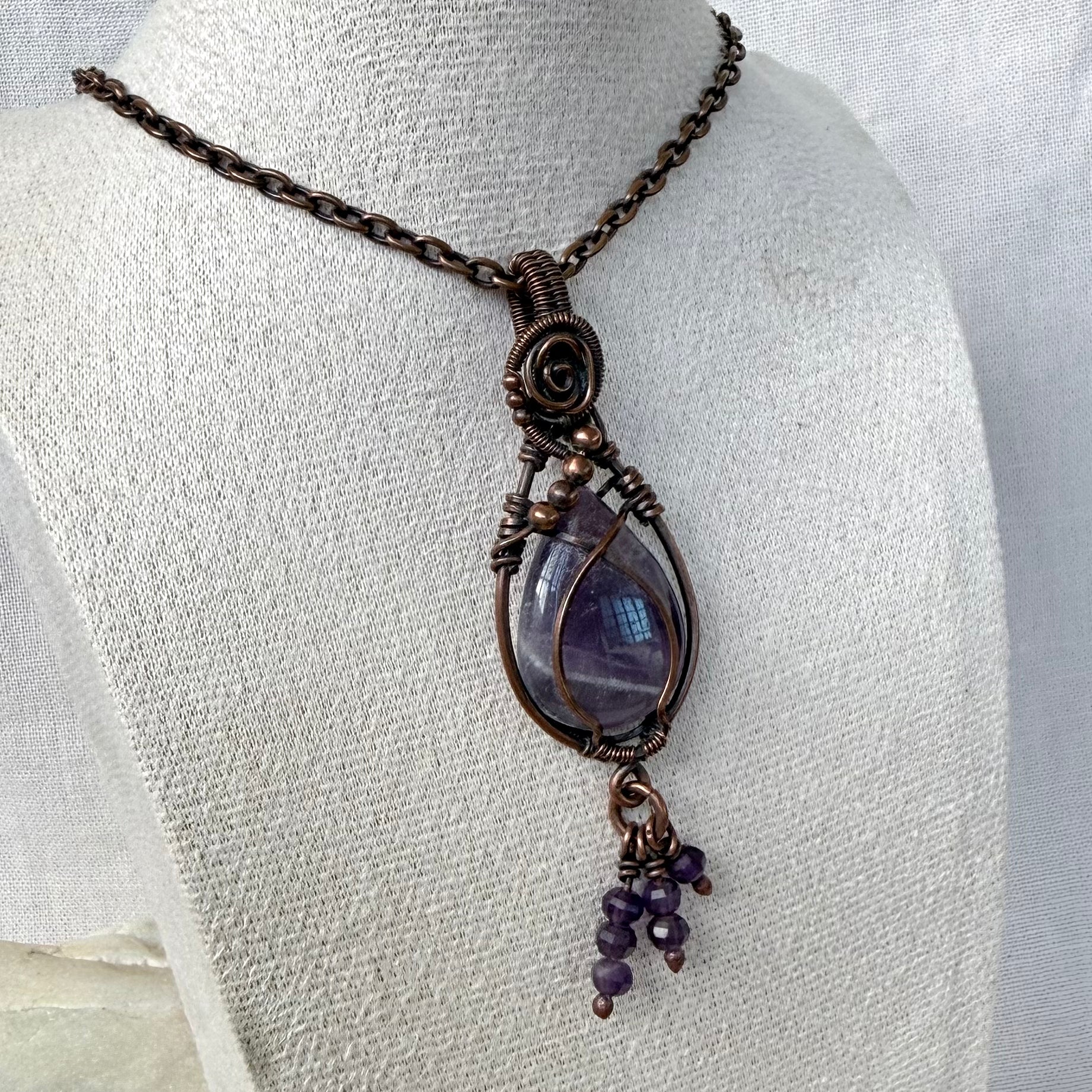 A teardrop-shaped amethyst pendant necklace with a purple stone, small purple amethyst beads hanging from the bottom and copper wire wrap, displayed on a textured white fabric mannequin bust.