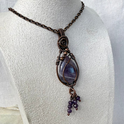 A teardrop-shaped amethyst pendant necklace with a purple stone, small purple amethyst beads hanging from the bottom and copper wire wrap, displayed on a textured white fabric mannequin bust.
