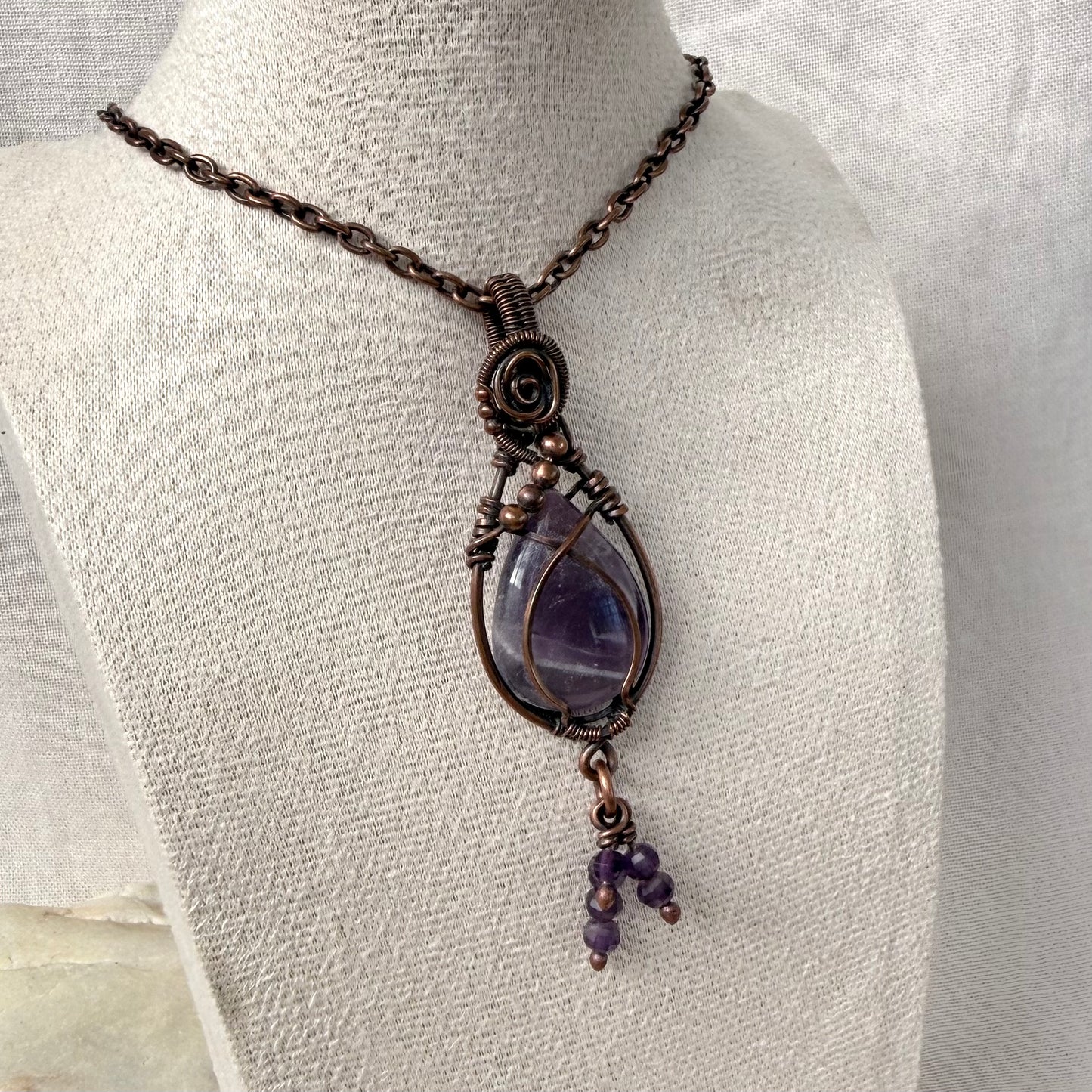 A handmade copper wire-wrapped amethyst pendant necklace displayed on a textured cream fabric mannequin bust. The necklace features a dark copper-toned metal chain and a central teardrop-shaped purple amethyst stone wrapped in detailed copper-toned wire, with a small dangling amethyst bead cluster at the bottom.