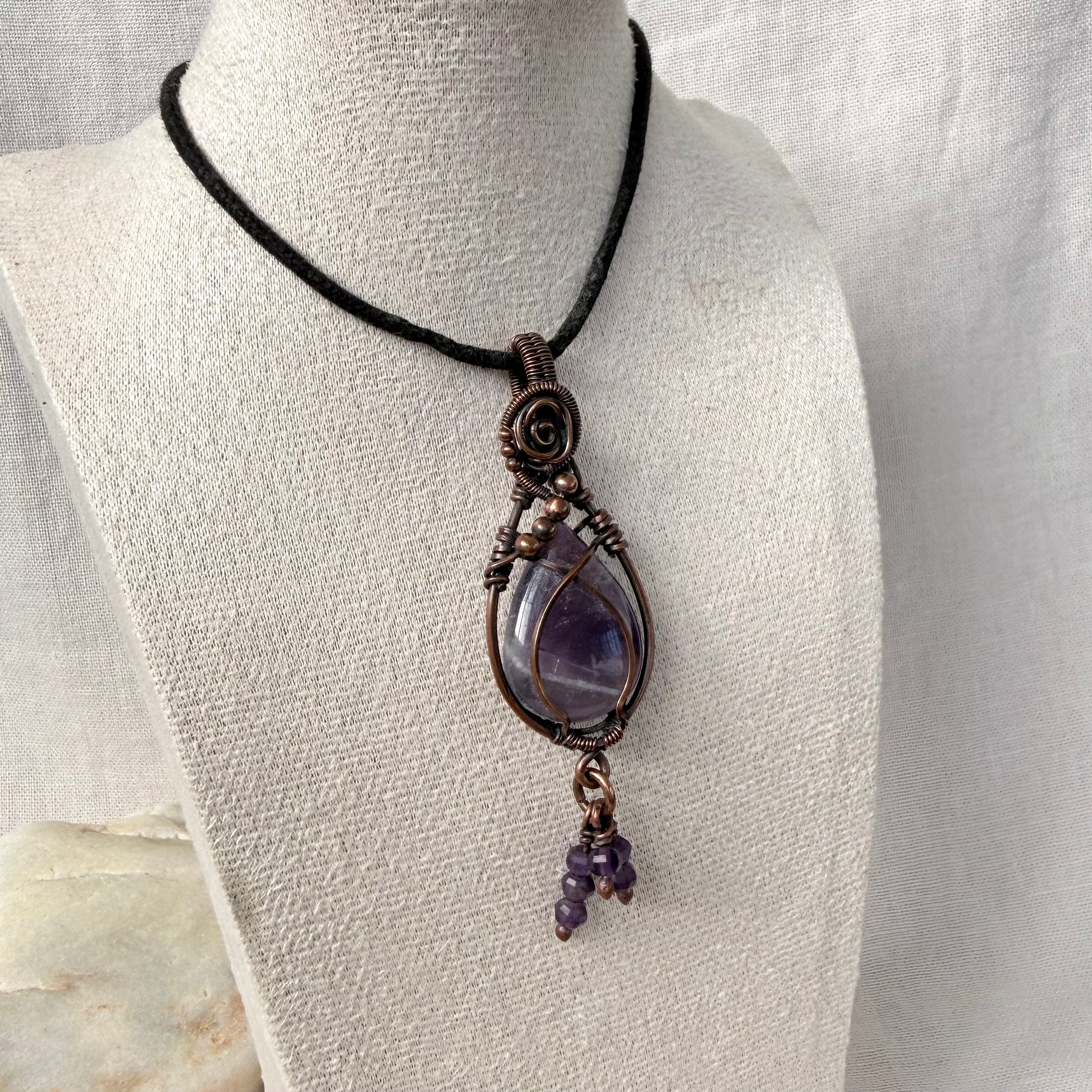 A handmade wire-wrapped amethyst pendant necklace on a black cord, displayed on a textured off-white fabric mannequin bust.