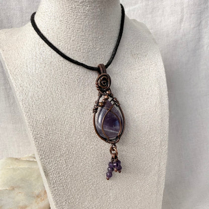 A handmade wire-wrapped amethyst pendant necklace on a black cord, displayed on a textured off-white fabric mannequin bust.