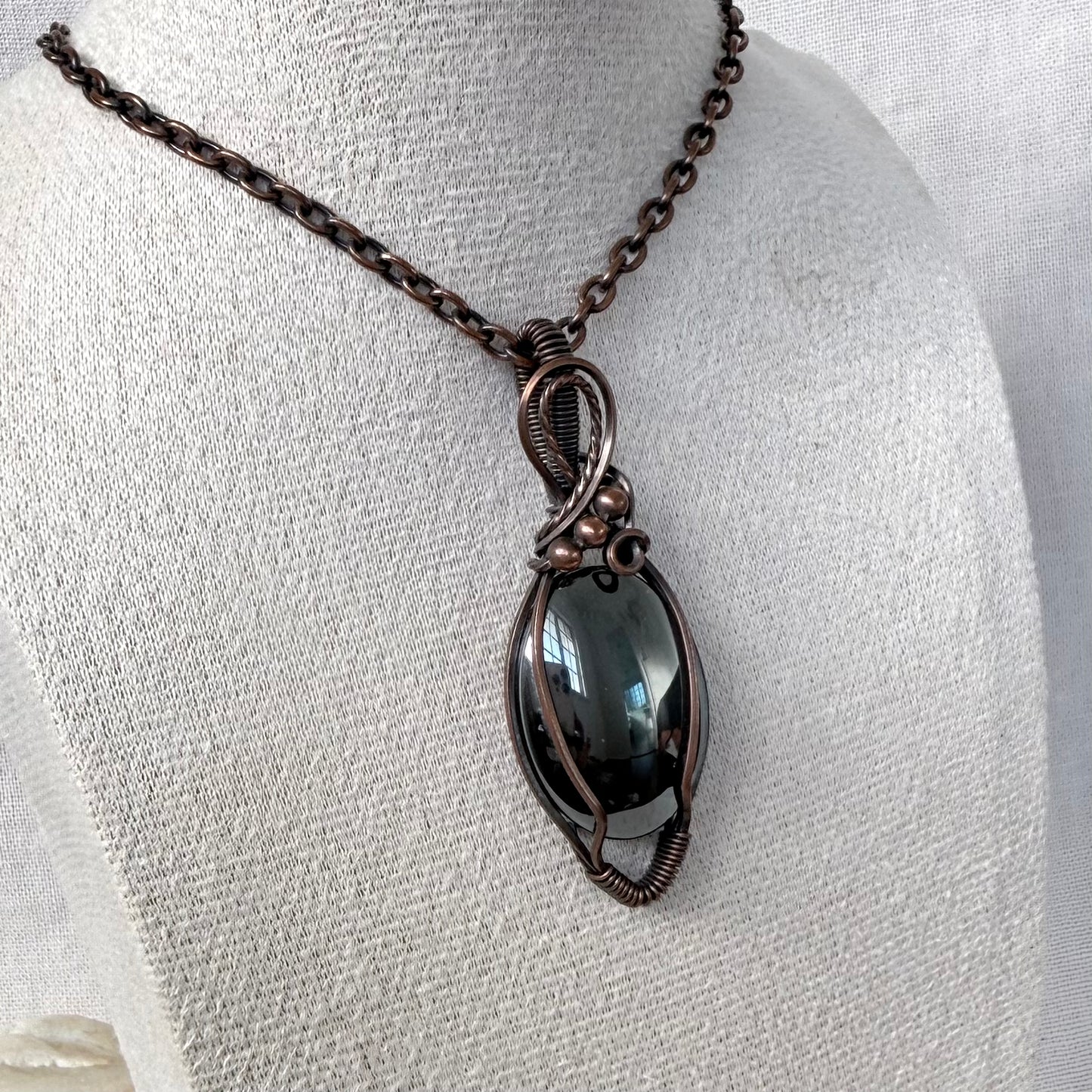 A dark smoky Hematite pendant necklace wrapped in a copper wire frame, displayed on a textured off-white fabric mannequin bust.