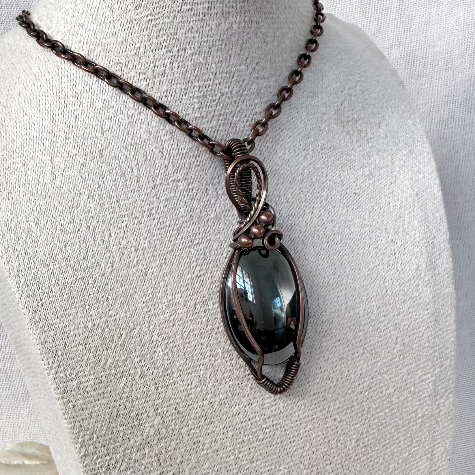 A dark smoky Hematite pendant necklace wrapped in a copper wire frame, displayed on a textured off-white fabric mannequin bust.