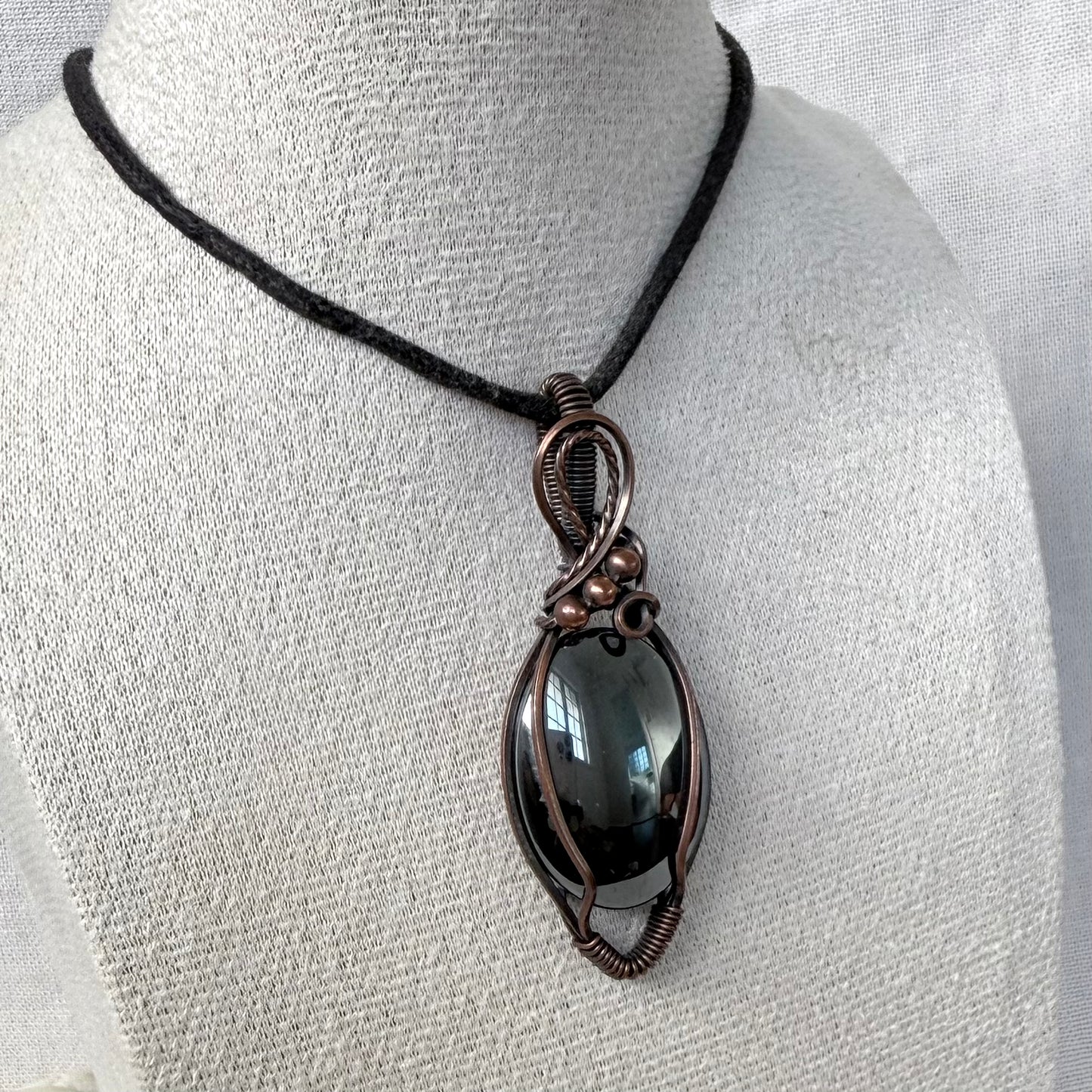 A handmade oval pendant necklace with a dark smoky Hematite stone wrapped in copper wire, displayed on a textured white mannequin bust with a black cord.
