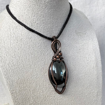 A handmade oval pendant necklace with a dark smoky Hematite stone wrapped in copper wire, displayed on a textured white mannequin bust with a black cord.