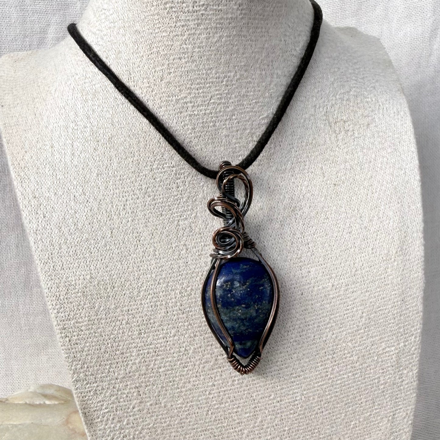 A teardrop-shaped lapis lazuli pendant necklace with a dark blue stone and copper wire wrap, displayed on a textured white fabric mannequin bust.