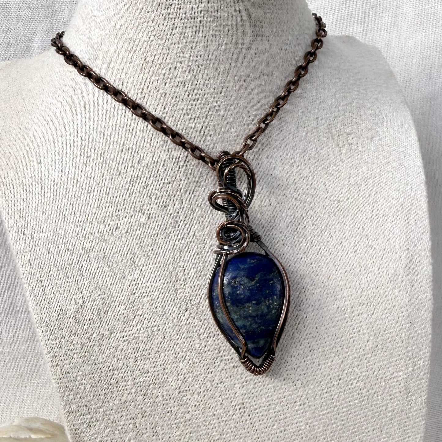 A copper teardrop-shaped lapis lazuli pendant necklace with a dark oxidized finish, wrapped in matching wire, displayed on a textured white fabric mannequin bust.