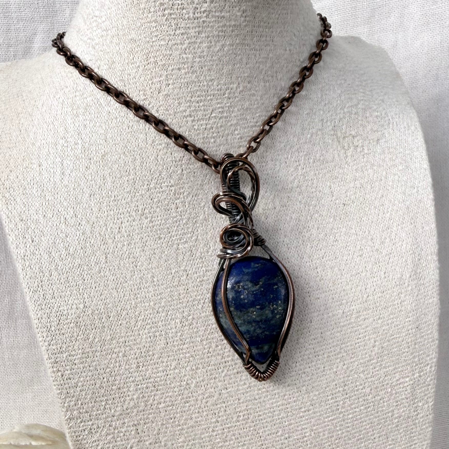 A copper teardrop-shaped lapis lazuli pendant necklace with a dark oxidized finish, wrapped in matching wire, displayed on a textured white fabric mannequin bust.