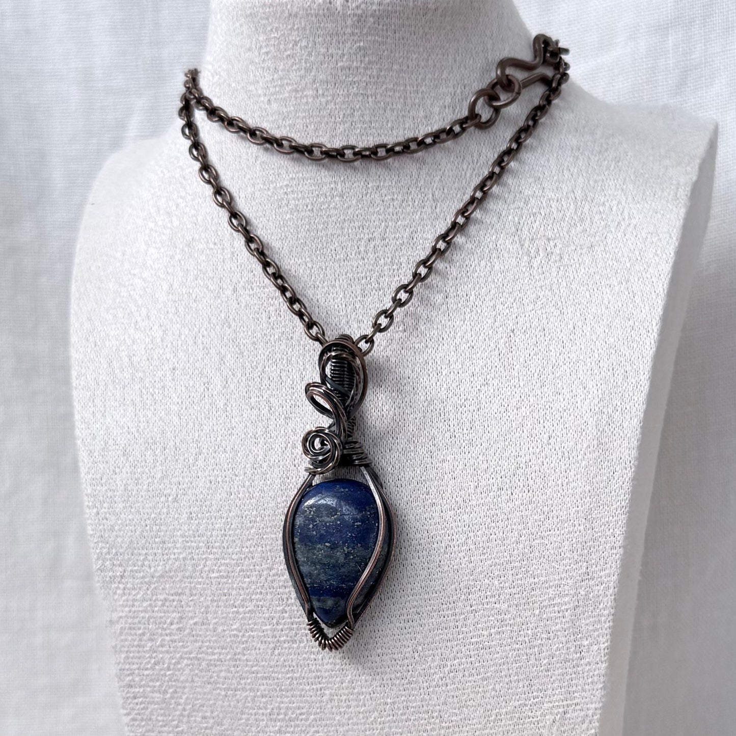 A dark oxidized copper necklace displayed on a white fabric mannequin bust. The necklace features a dark copper chain and a teardrop-shaped pendant with a natural blue lapis lazuli stone wrapped in dark metal.