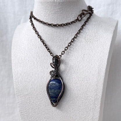 A dark oxidized copper necklace displayed on a white fabric mannequin bust. The necklace features a dark copper chain and a teardrop-shaped pendant with a natural blue lapis lazuli stone wrapped in dark metal.