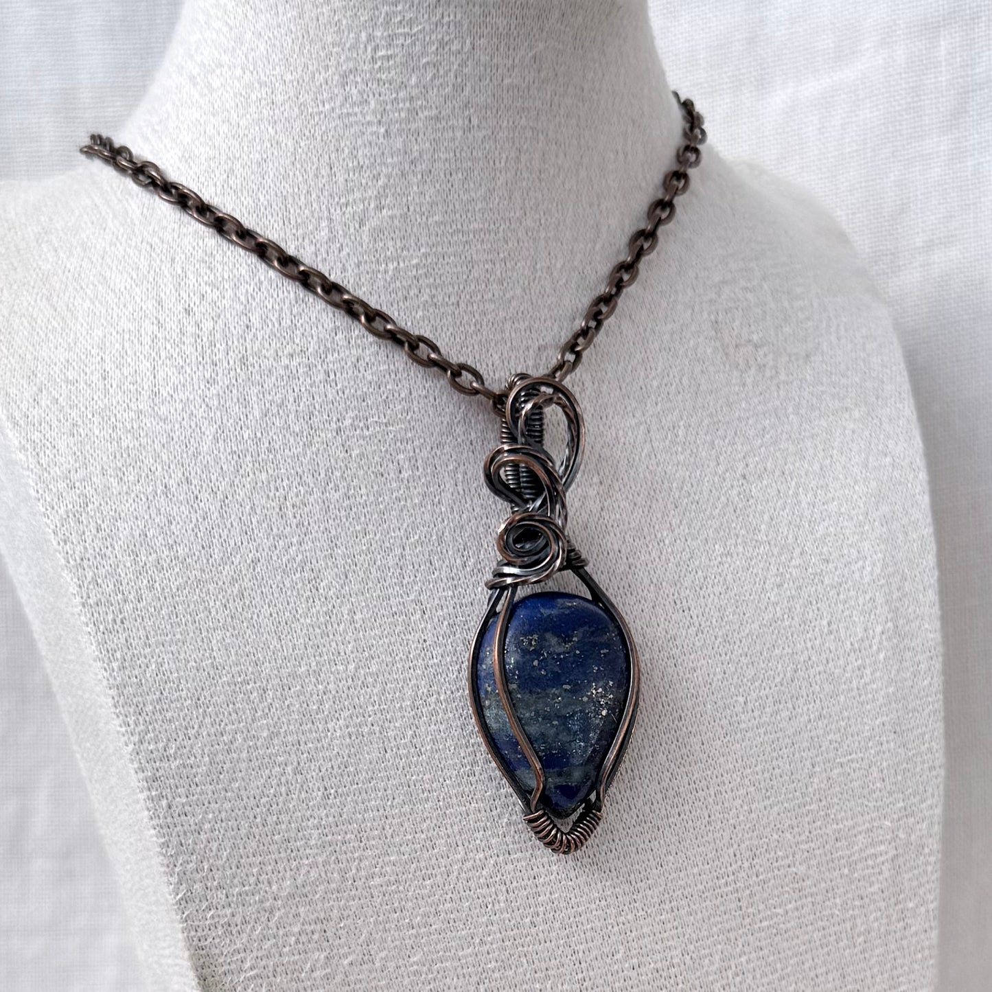 A handmade copper wire-wrapped pendant necklace featuring a blue lapis lazuli stone with gold veining, displayed on a white fabric mannequin bust.