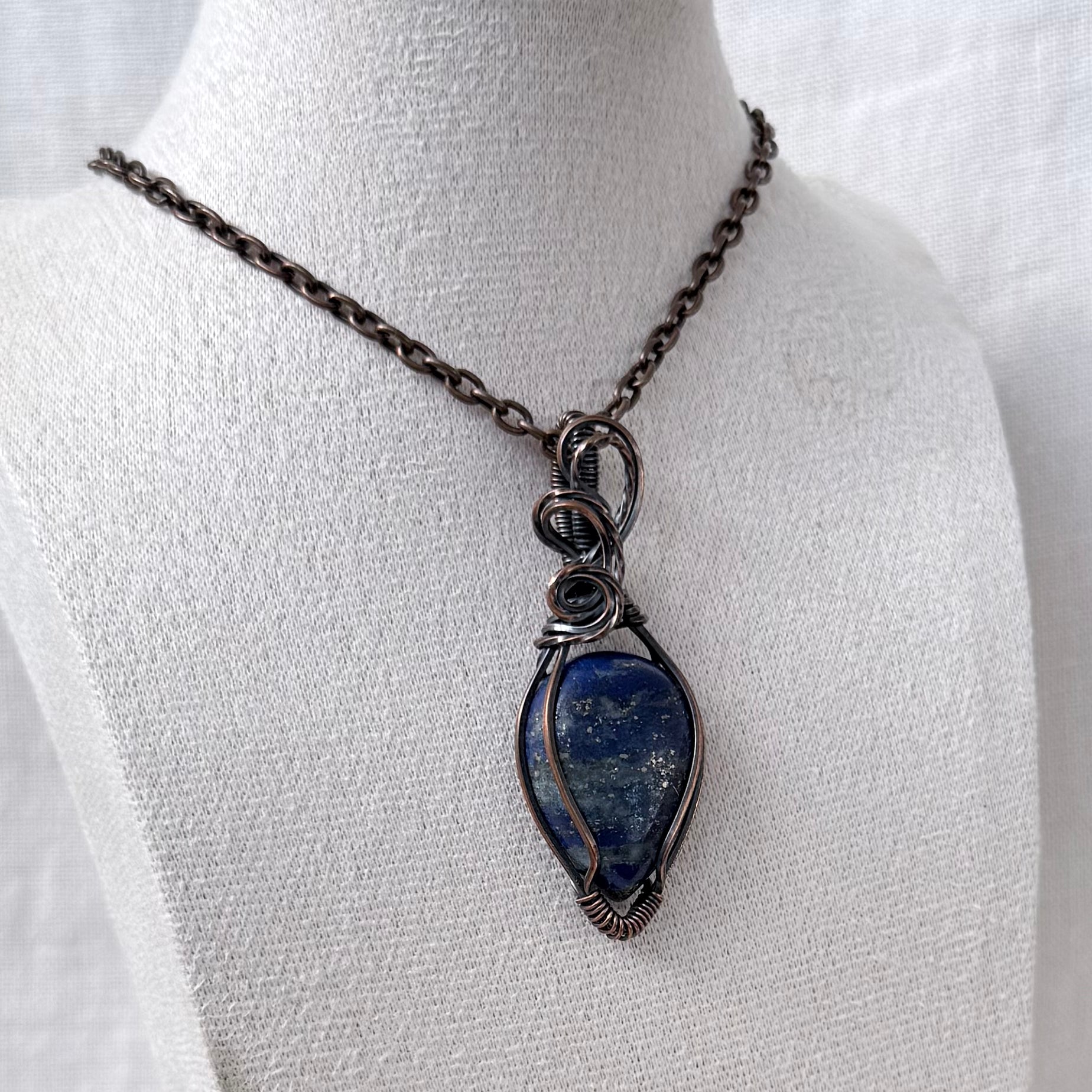 A handmade copper wire-wrapped pendant necklace featuring a blue lapis lazuli stone with gold veining, displayed on a white fabric mannequin bust.