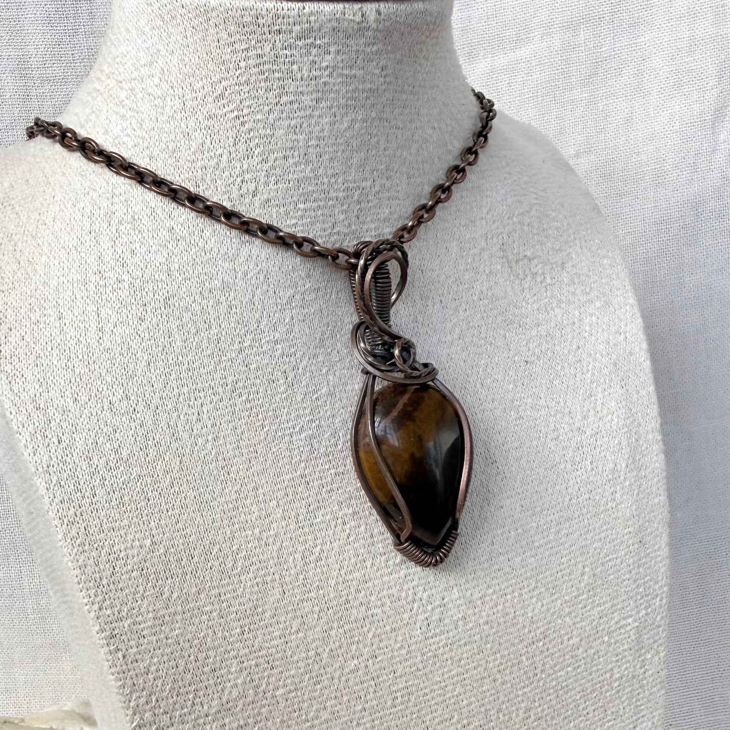 A handmade wire-wrapped pendant necklace featuring a polished brown tiger's eye stone in a copper wire setting, displayed on a textured off-white fabric mannequin bust.