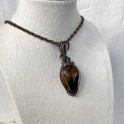 A handmade wire-wrapped pendant necklace featuring a polished brown tiger's eye stone in a copper wire setting, displayed on a textured off-white fabric mannequin bust.
