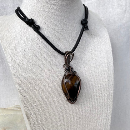 A handmade necklace featuring a polished brown tiger's eye stone wrapped in dark copper wire, suspended from a black cord with knot details, displayed on a textured white fabric mannequin bust.