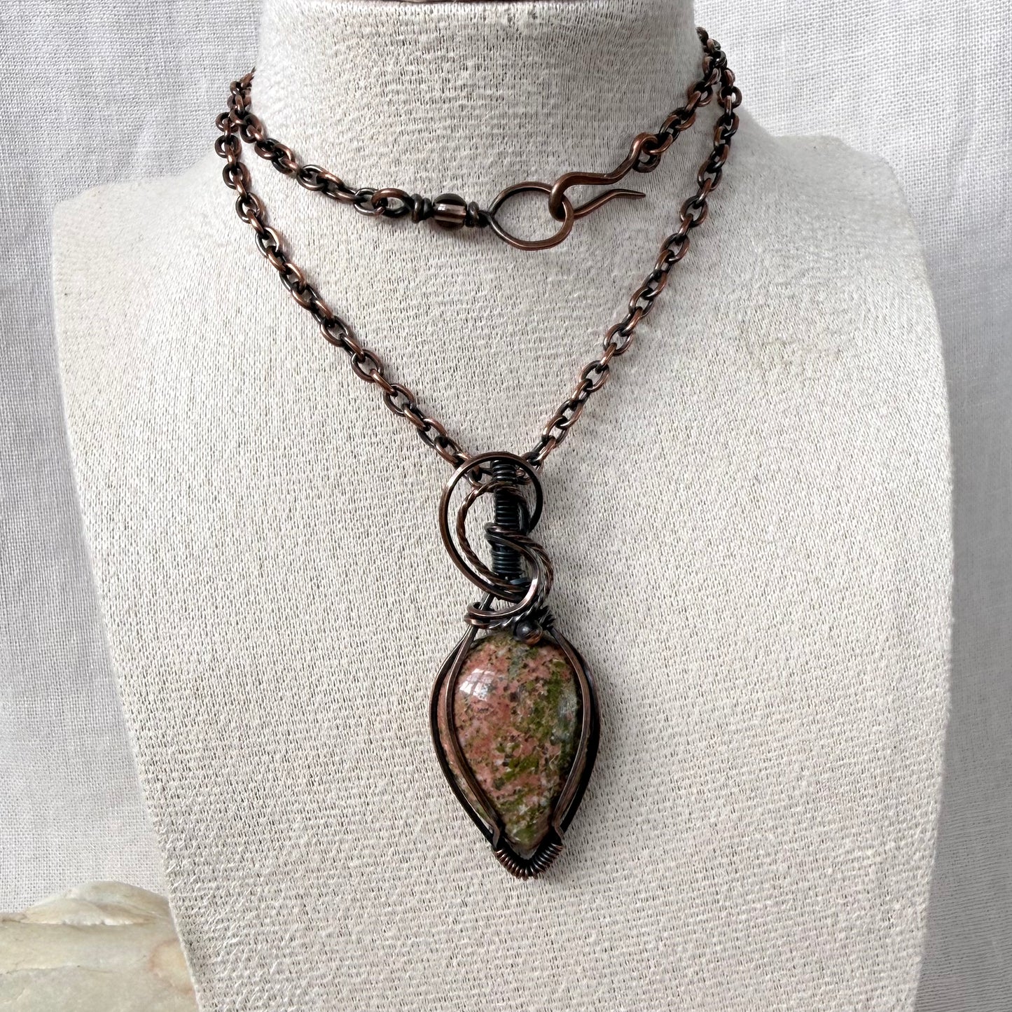 A copper necklace displayed on a cream fabric mannequin bust. The necklace features copper chains with a hook clasp on the top strand and a larger teardrop-shaped pendant with a Unakite stone on the bottom.