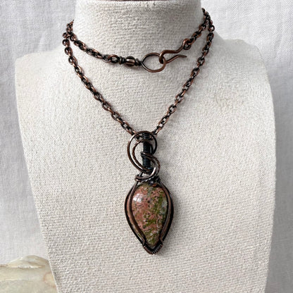 A copper necklace displayed on a cream fabric mannequin bust. The necklace features copper chains with a hook clasp on the top strand and a larger teardrop-shaped pendant with a Unakite stone on the bottom.