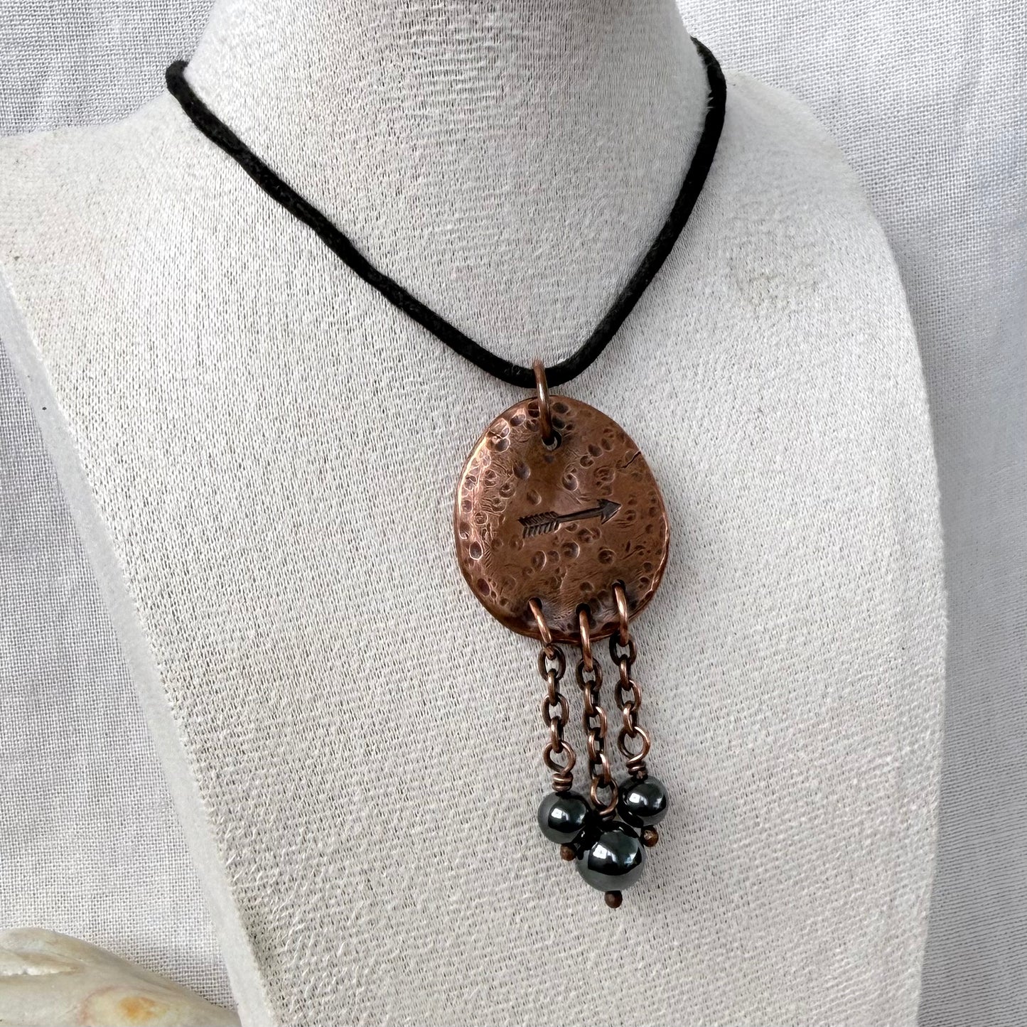A handmade bohemian-style copper pendant necklace displayed on a white fabric mannequin bust. The necklace features a black cord, a hammered copper oval pendant with a small arrow engraving, and three dangling black bead chains.