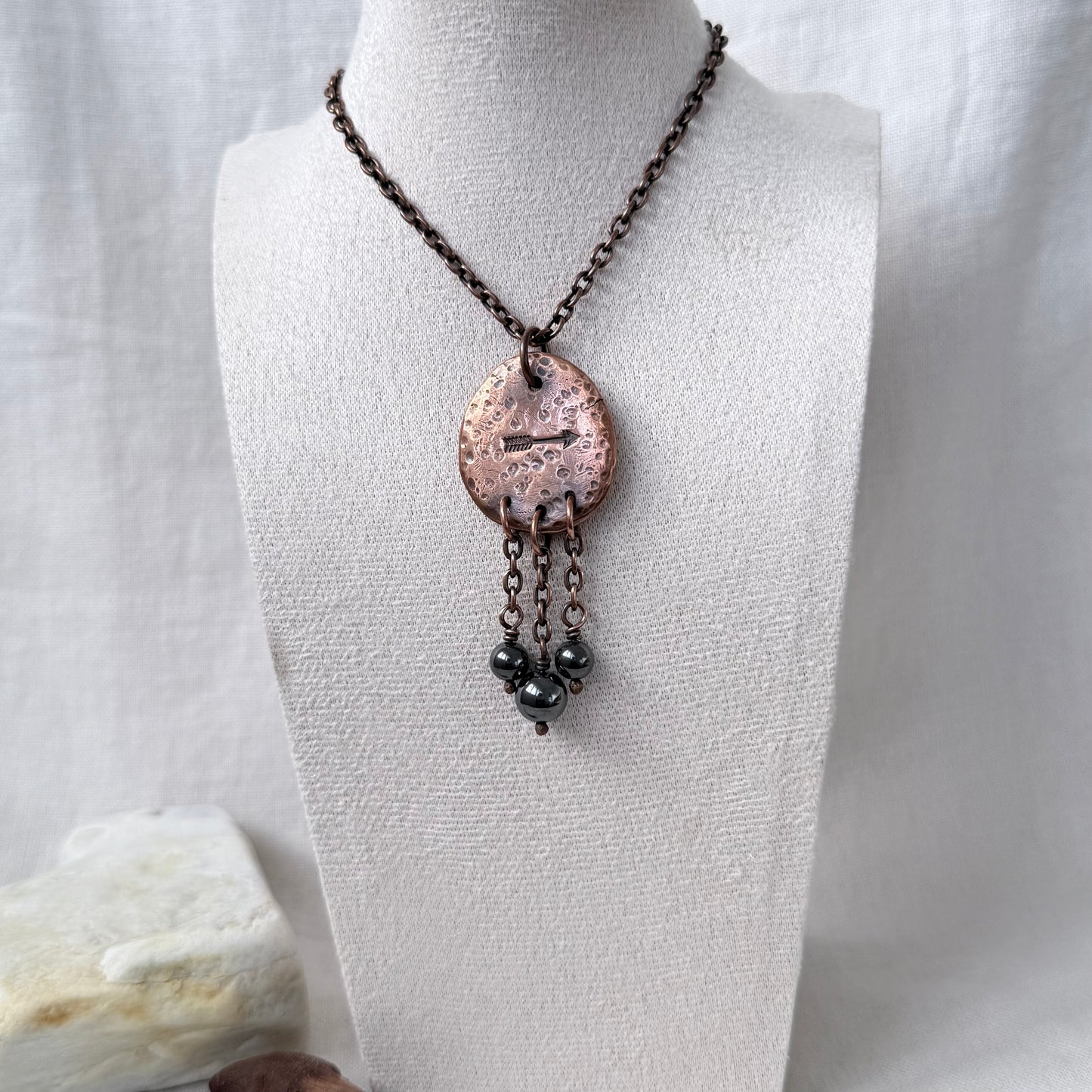 A handmade pendant necklace displayed on a white fabric mannequin bust. The necklace features a dark copper chain, a hammered copper oval pendant, and three dangling dark grey beads.