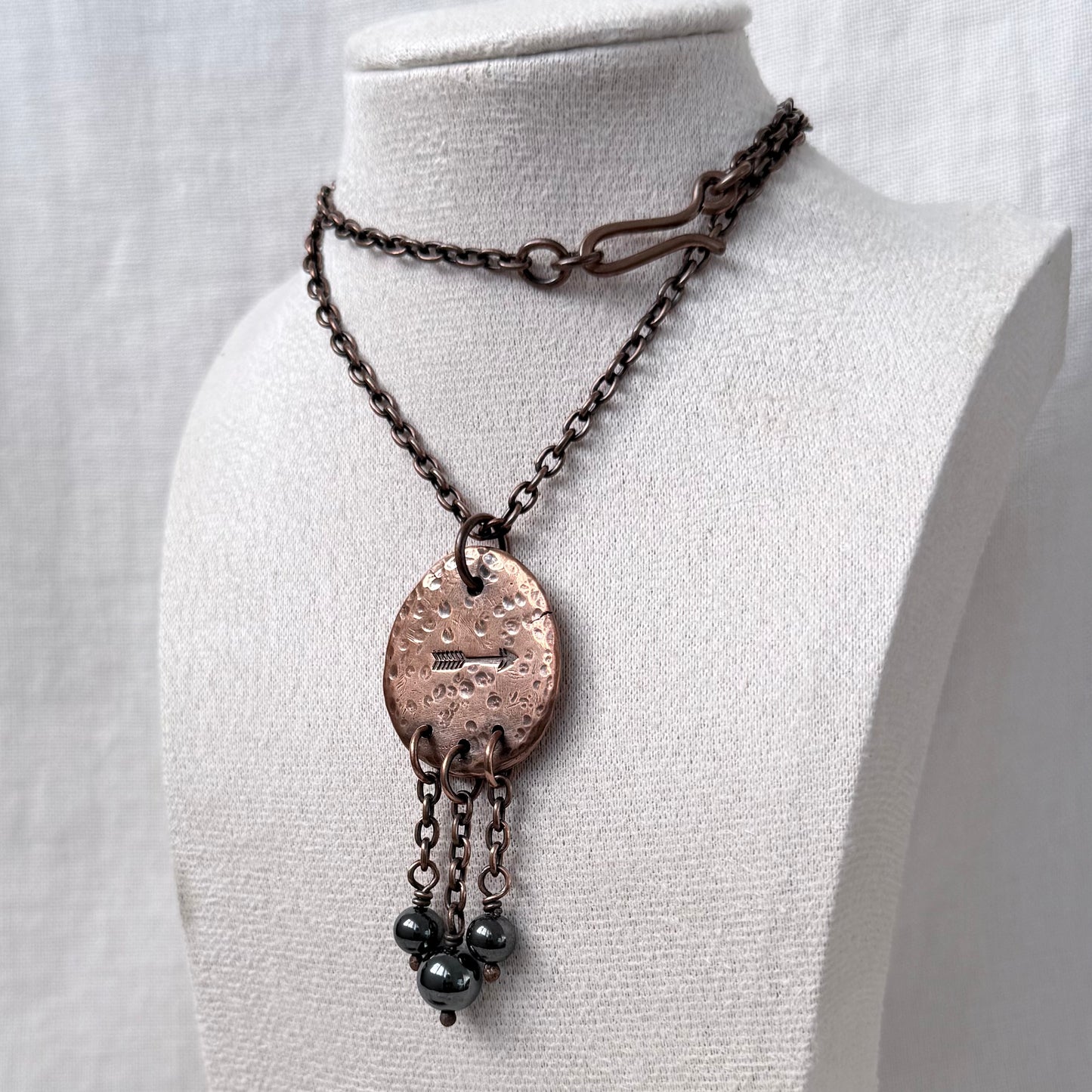 A handmade pendant necklace displayed on a white fabric mannequin bust. The necklace features a dark copper chain, a hammered copper oval pendant, and three dangling dark grey beads.