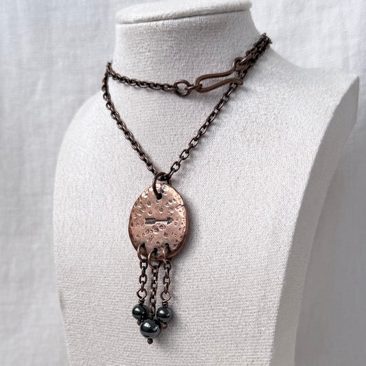 A handmade pendant necklace displayed on a white fabric mannequin bust. The necklace features a dark copper chain, a hammered copper oval pendant, and three dangling dark grey beads.