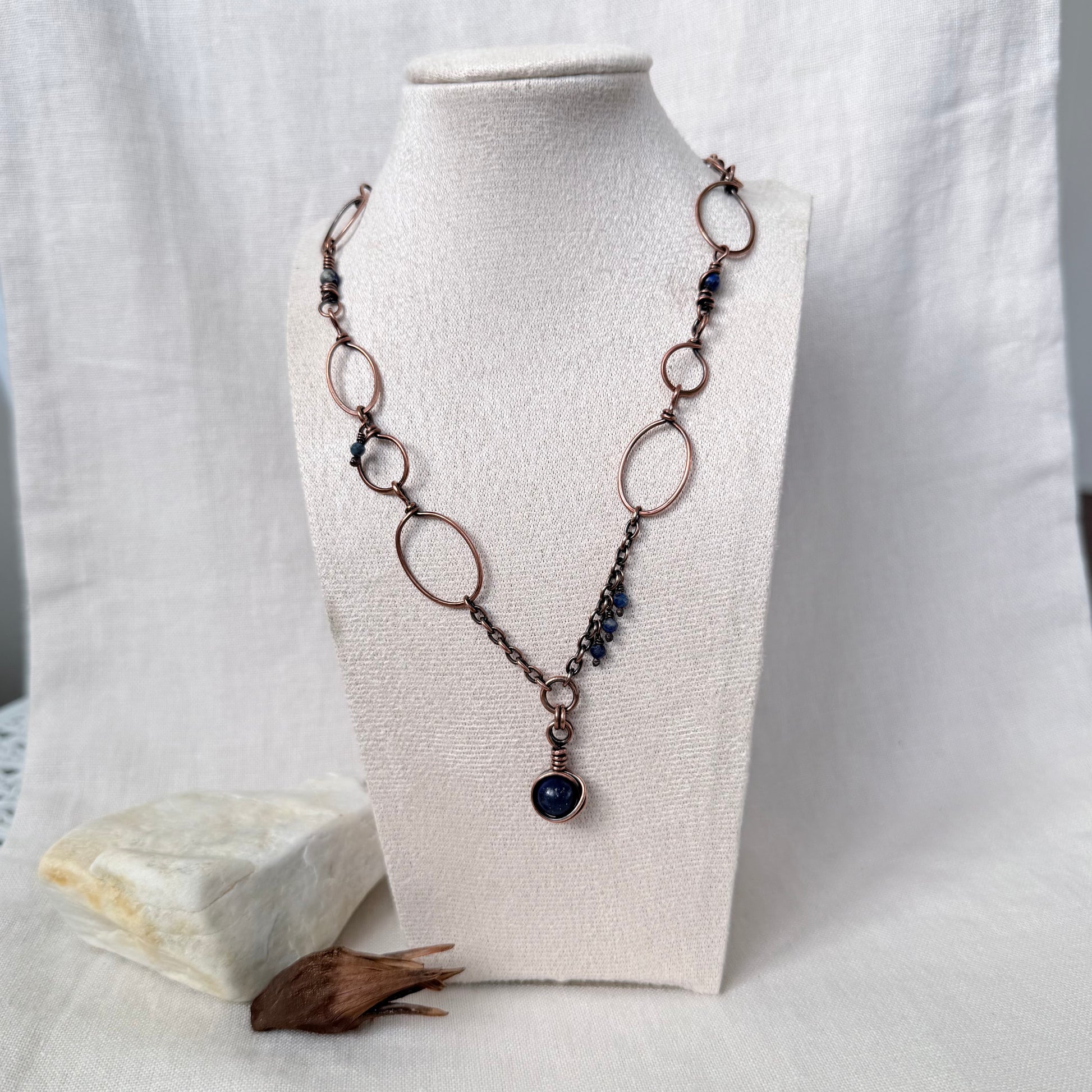 A handmade copper necklace with oval links, small dark beads, and a dark blue stone pendant, displayed on a cream fabric mannequin bust.