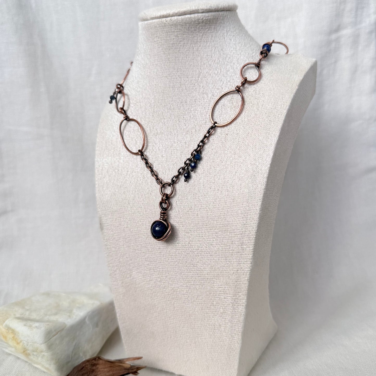 A handmade copper and dark metal chain necklace with a dark blue stone pendant, displayed on a cream fabric-covered jewelry bust.