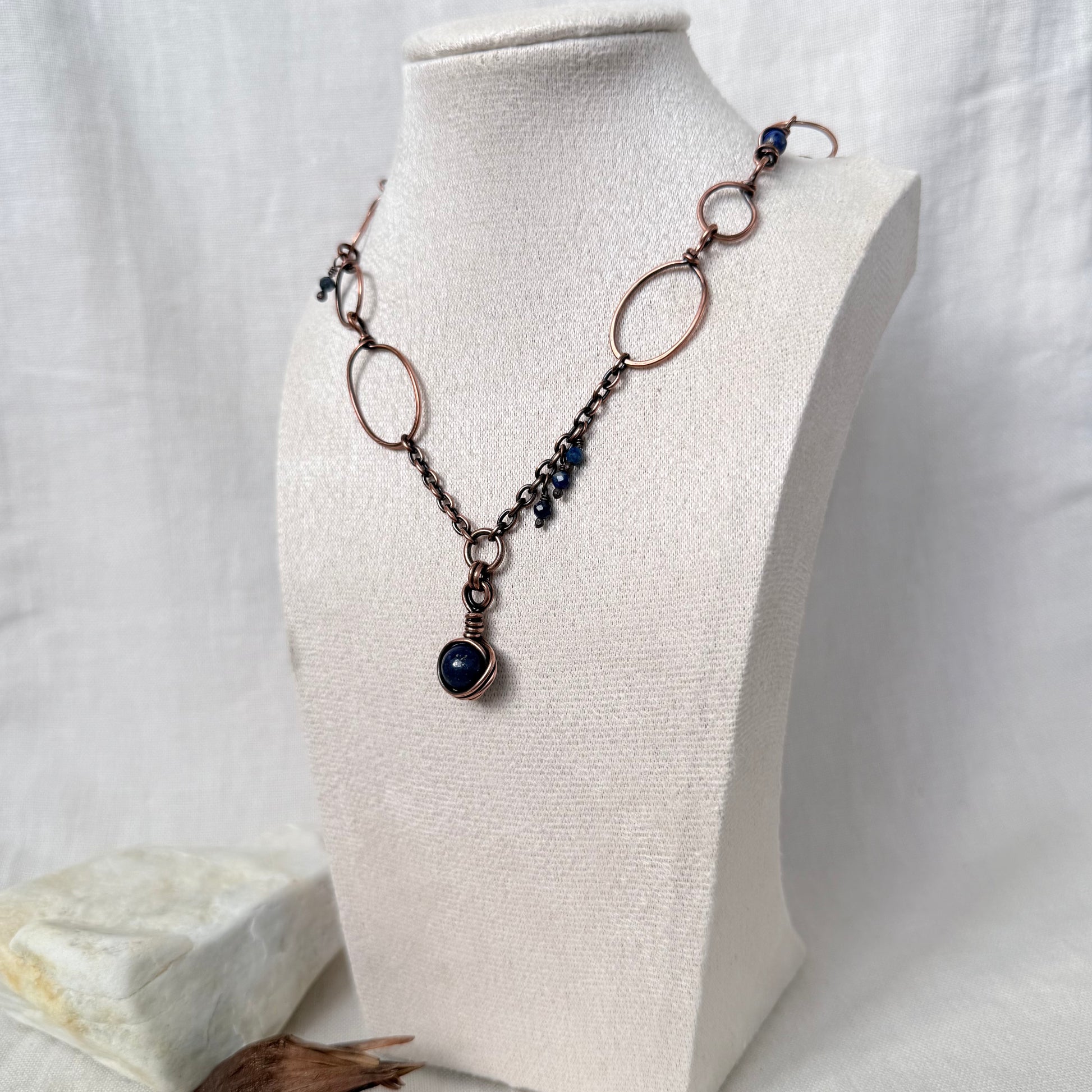 A handmade copper and dark metal chain necklace with a dark blue stone pendant, displayed on a cream fabric-covered jewelry bust.