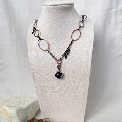 A handmade necklace with copper wire links, dark blue beads, and a central dark blue stone pendant, displayed on a cream fabric mannequin bust.