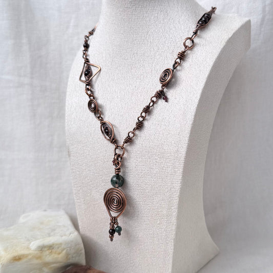 A copper bohemian-style necklace displayed on a white fabric mannequin bust. The necklace features multiple spiral wire details, small dark beads, and a larger dark green bead near the pendant.