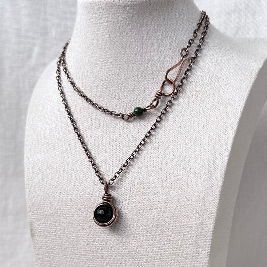 A copper necklace displayed on a white fabric mannequin bust. The necklace features a dark oxidized copper chain with a circular dark green Moss Agate stone pendant and a smaller green bead accent on the clasp.