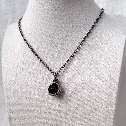 A necklace with a dark round Moss Agate stone in a copper wrap, displayed on a white fabric jewellery bust.