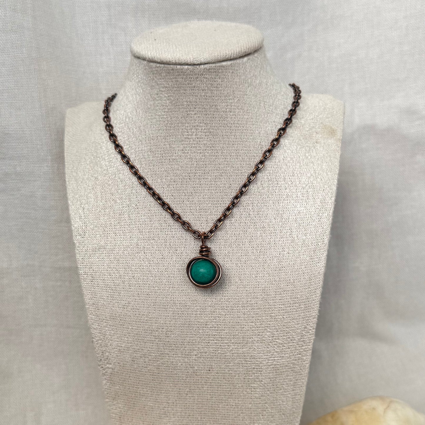 A copper necklace with a copper chain and a round pendant featuring a turquoise round gemstone, displayed on a textured beige fabric jewellery bust.